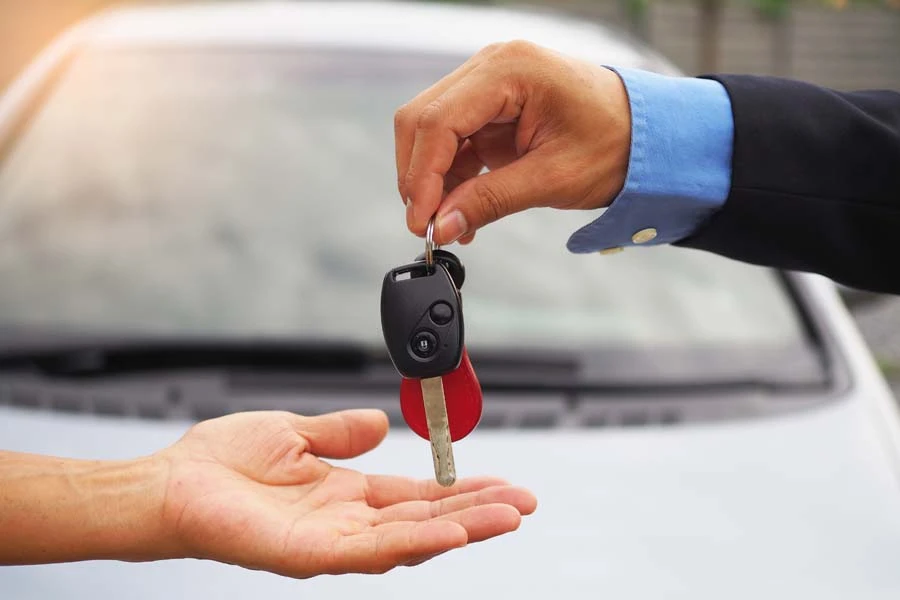 Une main en costume bleu marine transmet des cl&eacute;s de voiture &agrave; une main tendue, dans un environnement ext&eacute;rieur avec v&eacute;hicule blanc en arri&egrave;re-plan